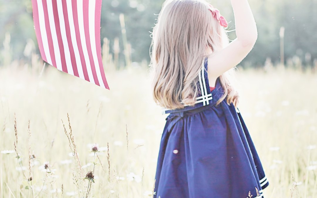 A young girl in a navy dress holding a US flag in a summer meadow
