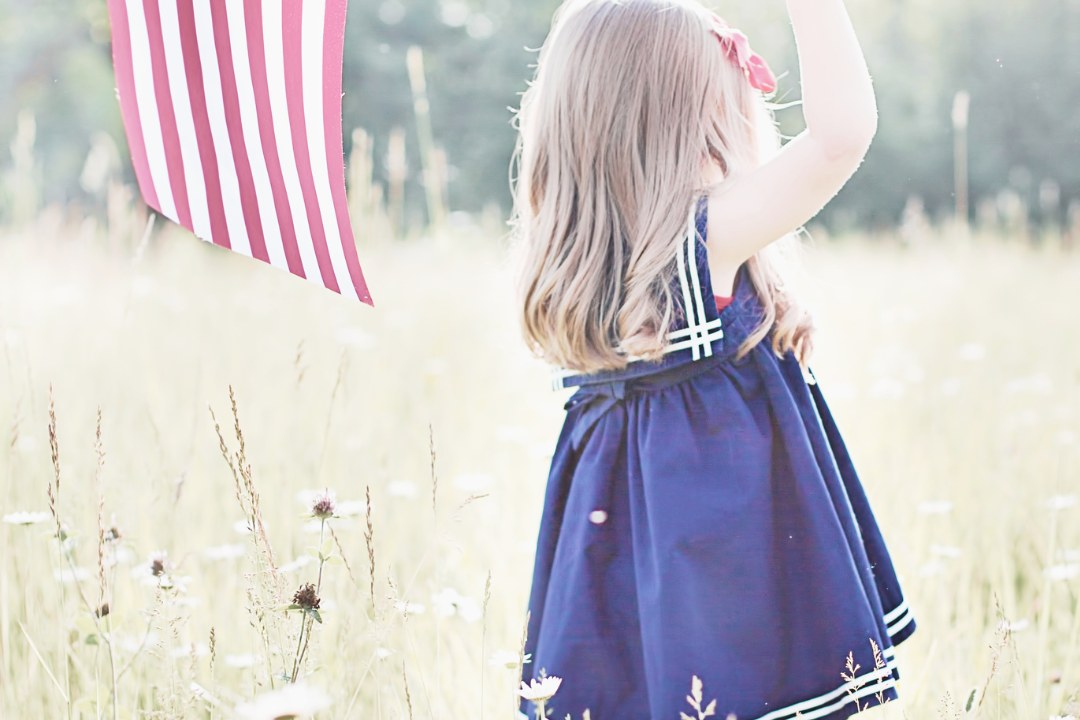 A young girl in a navy dress holding a US flag in a summer meadow